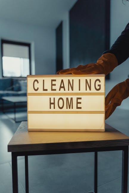 A close-up of a person wearing orange cleaning gloves, placing a wooden letterboard on a dark wooden table in a modern, well-lit room with large windows and minimal decor. The letterboard displays the